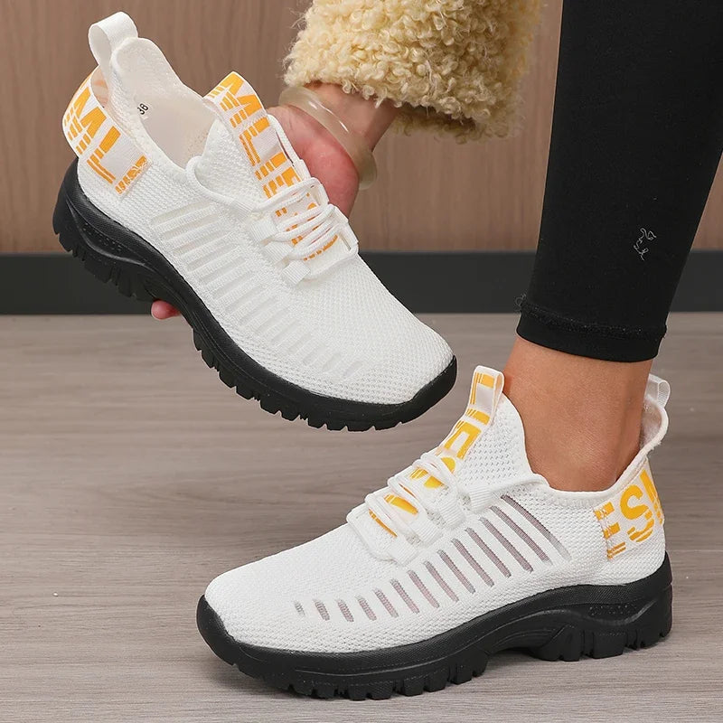 White sneakers with black soles and yellow accents worn by a person on a wooden floor.