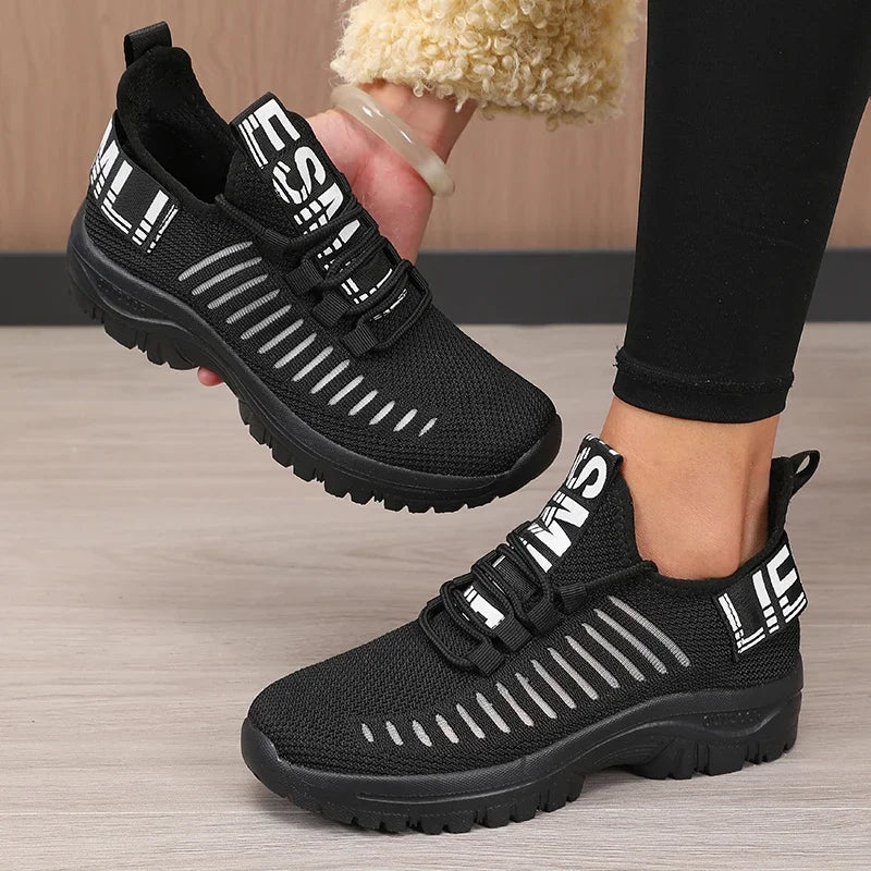 Black athletic shoes with white text worn by a person on a wooden floor.