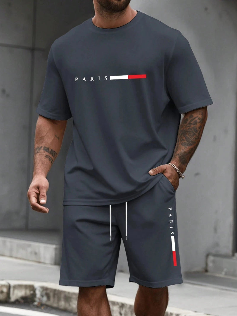 Person wearing a navy blue t-shirt and shorts with 'PARIS' logo, standing against a gray wall.