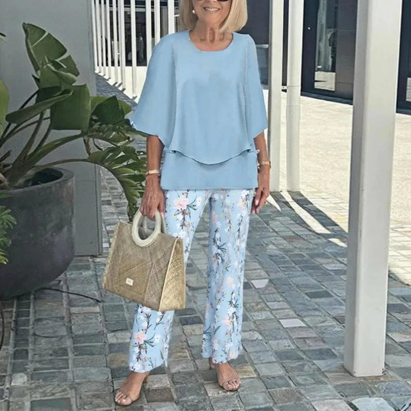 Woman in light blue outfit with floral pants holding a beige bag outdoors.