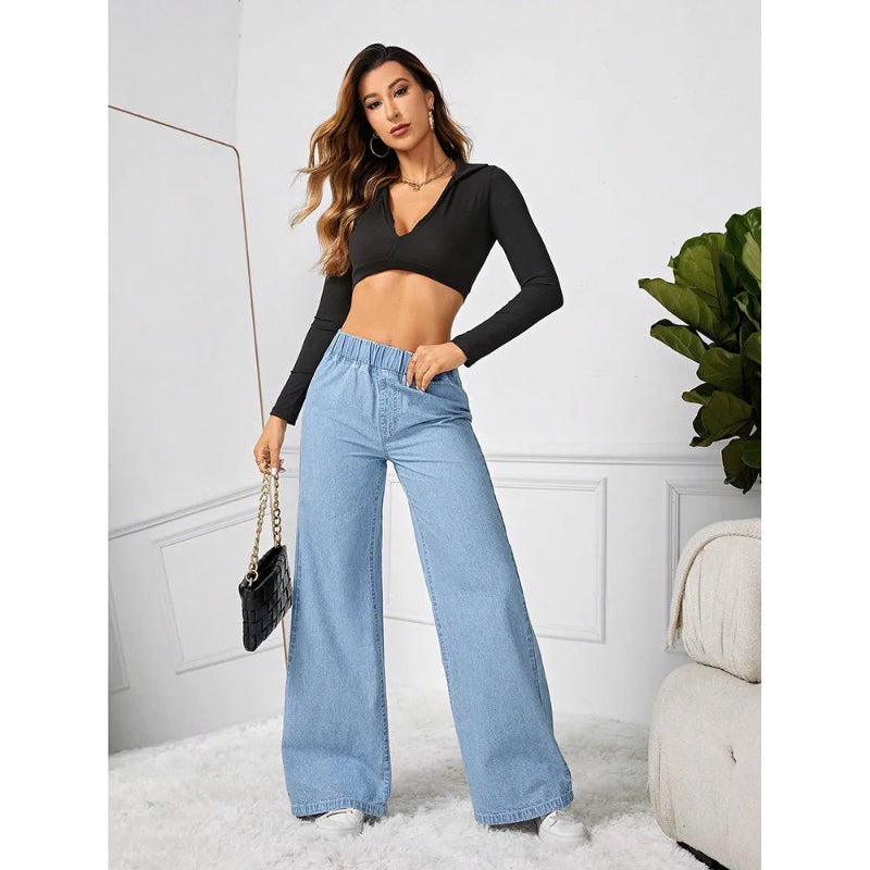 Woman wearing a black long-sleeve top and light blue jeans in a room with white walls and a plant.