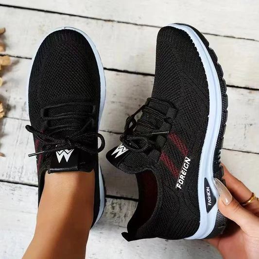 Black athletic shoes with white soles held by a person on a wooden floor.