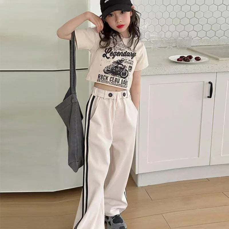 Person wearing a beige t-shirt with text and graphics, white pants with black stripes, and a black cap in a kitchen.