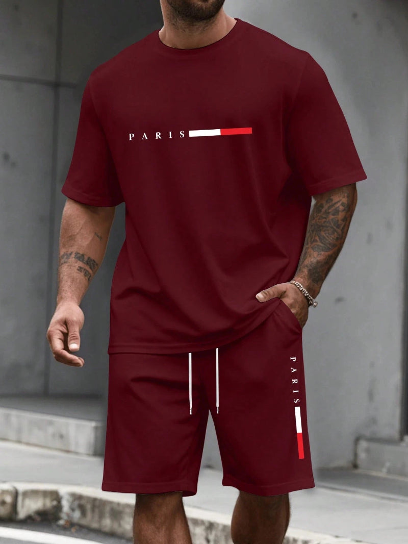 Man wearing a maroon 'PARIS' t-shirt and shorts with a concrete wall background