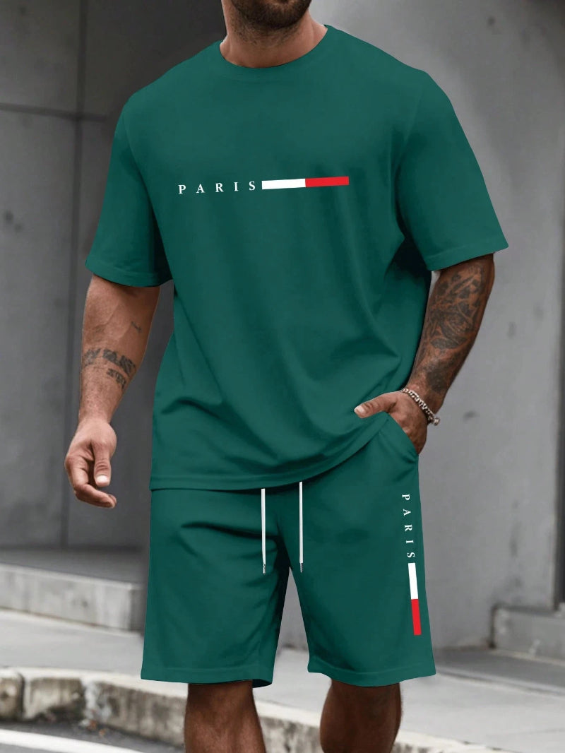Man wearing a green 'PARIS' t-shirt and shorts with a logo against a gray wall.