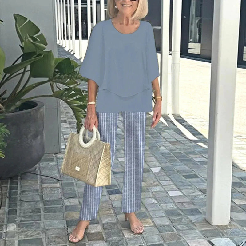 Woman in a blue outfit with striped pants holding a beige bag on a stone patio.
