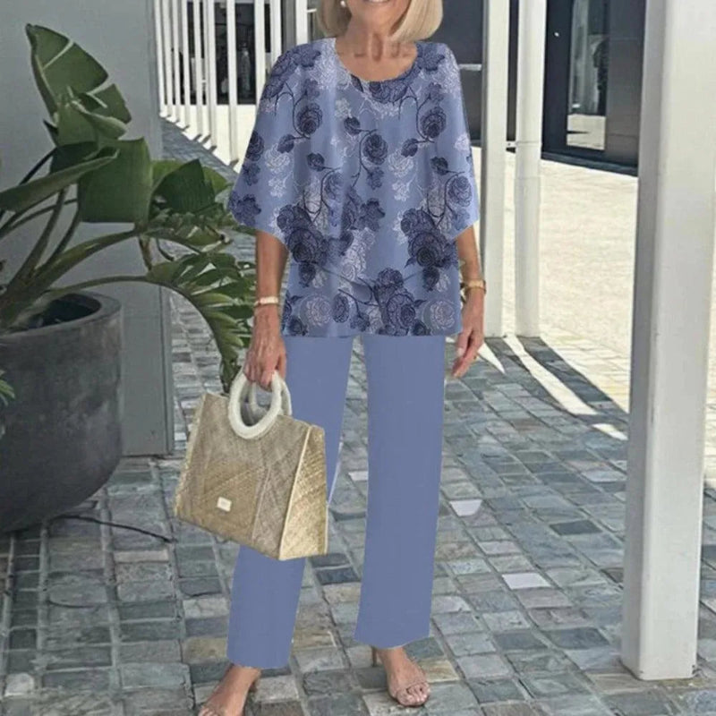 Woman in a blue floral top and pants holding a beige handbag on a stone patio.